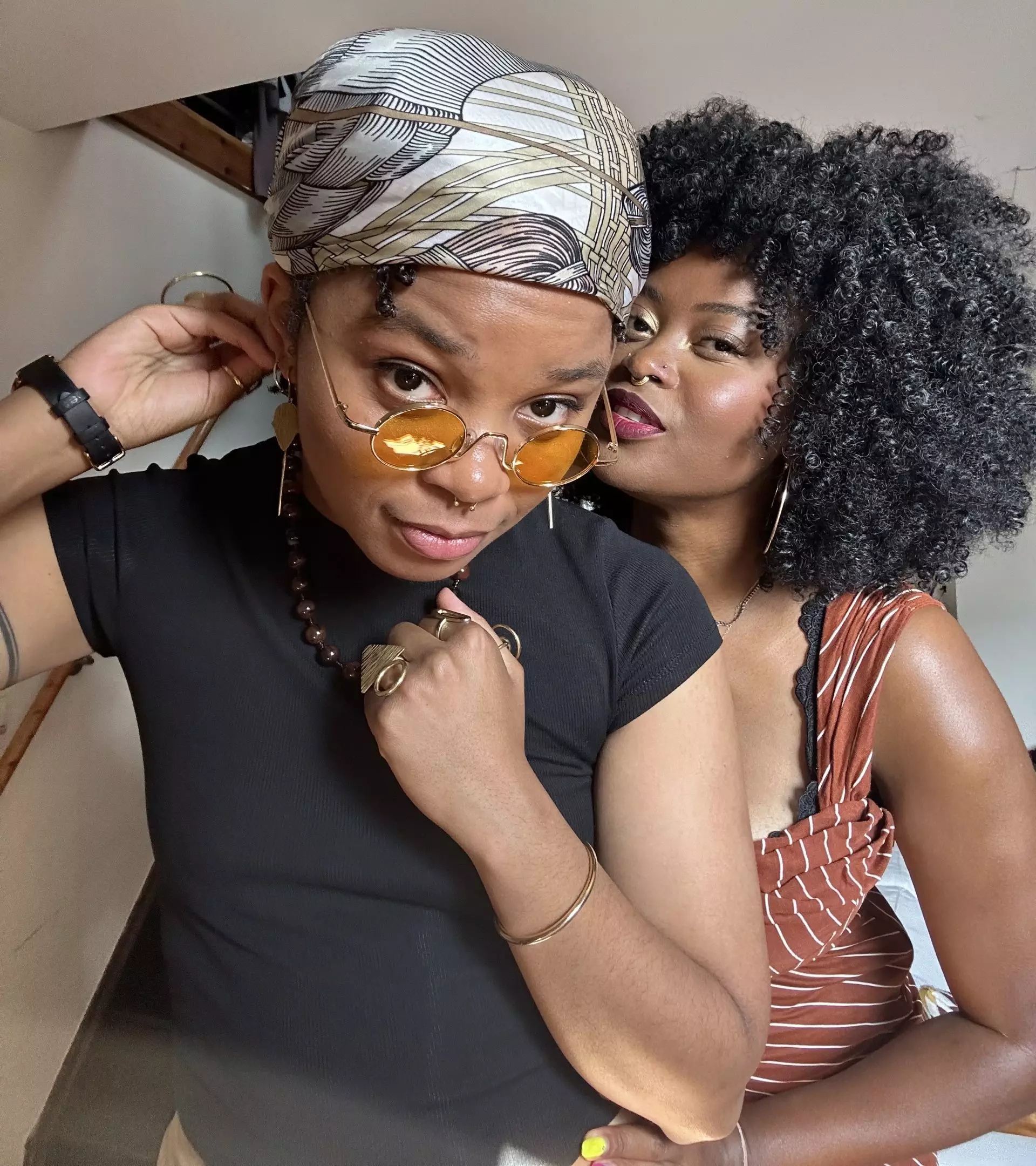 A couple of Black queer women. One is in the foreground sporting a silk beige scarf and the other is other is in the background peeking over the shoulder of the one infront. She has an afro and is wearing a brown and beige striped dress.