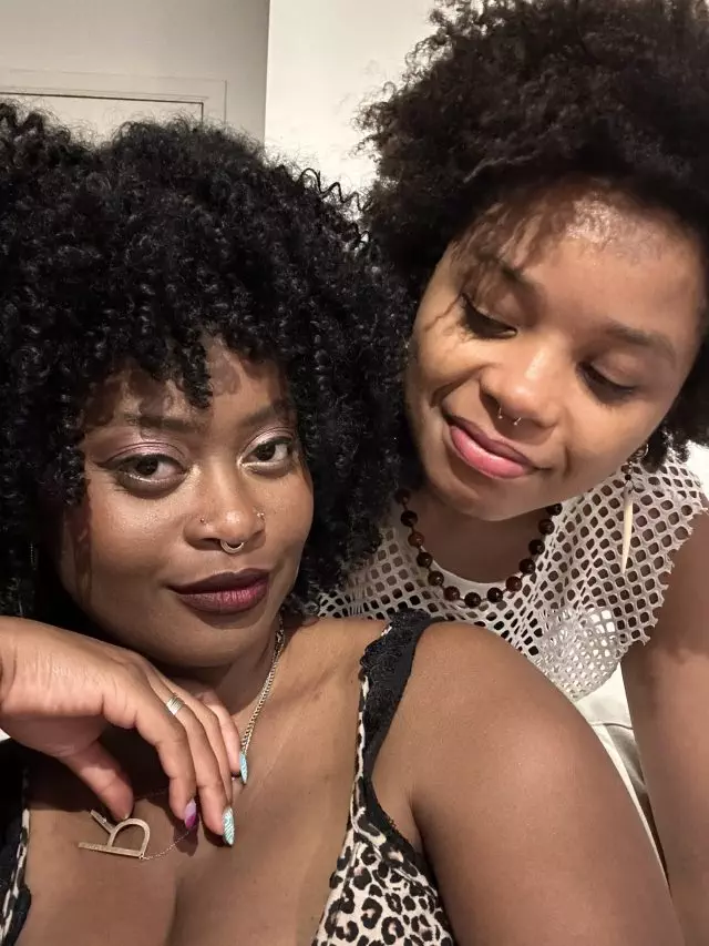A photo of RaeRae and Bo, two Black and queer women with afros. RaeRae is looking straight into the camera whilst Bo is placed behind her whilst leaning over and looking at RaeRae