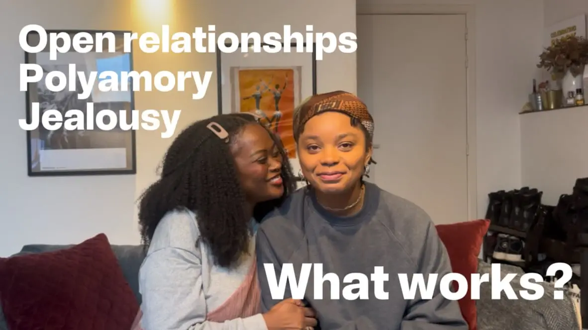 Two Black women, RaeRae and Bo, sitting close together and smiling at the camera against a neutral background. They're wearing casual attire as they discuss their polyamorous relationship journey.